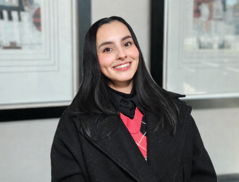Smiling woman in a black coat poses indoors with framed pictures behind her.